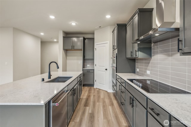 kitchen featuring wall chimney exhaust hood, stainless steel appliances, light wood finished floors, a sink, and light stone counters