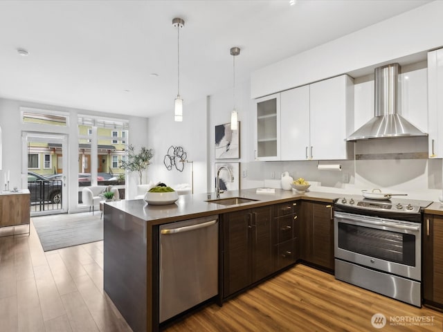 kitchen with appliances with stainless steel finishes, white cabinets, a sink, wood finished floors, and wall chimney exhaust hood