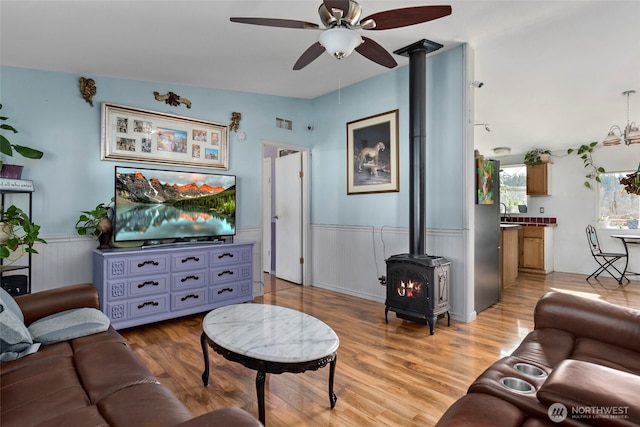 living area featuring light wood finished floors, visible vents, a wood stove, wainscoting, and ceiling fan