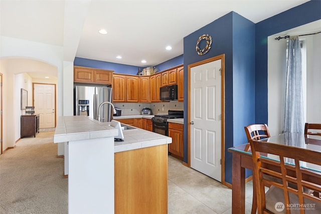 kitchen featuring tile countertops, decorative backsplash, a sink, an island with sink, and black appliances