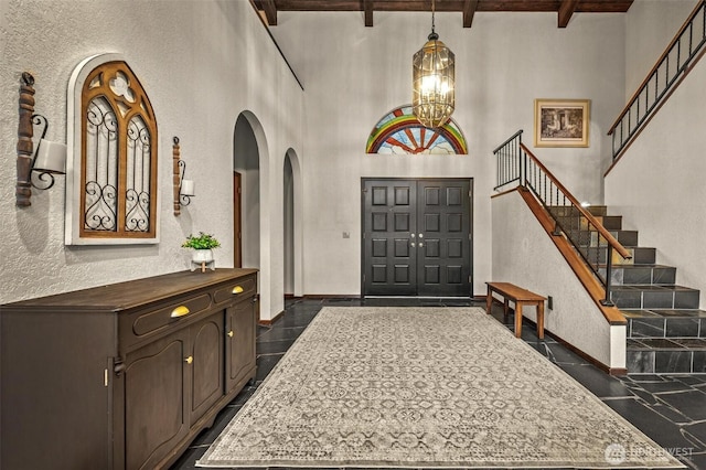 entryway featuring a notable chandelier, a textured wall, a towering ceiling, beamed ceiling, and stairs