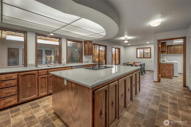 kitchen featuring light stone counters, black electric cooktop, a sink, a center island, and brown cabinetry