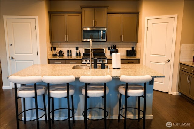 kitchen with light stone counters, dark wood-style flooring, stainless steel appliances, backsplash, and an island with sink