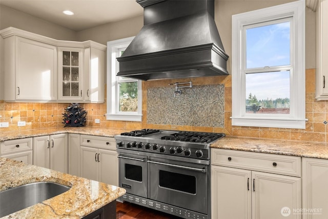 kitchen featuring range with two ovens, light stone counters, custom range hood, glass insert cabinets, and white cabinetry