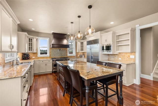 kitchen with a center island with sink, custom exhaust hood, open shelves, a sink, and built in appliances