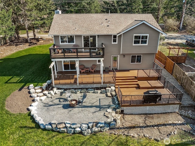 back of house featuring a deck, an outdoor fire pit, roof with shingles, a lawn, and a chimney