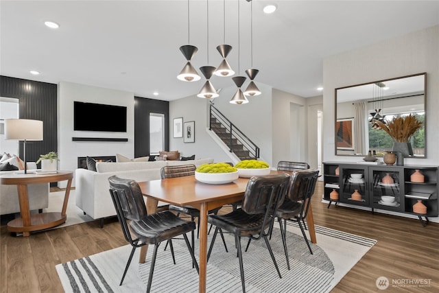 dining area featuring a warm lit fireplace, stairway, wood finished floors, and recessed lighting