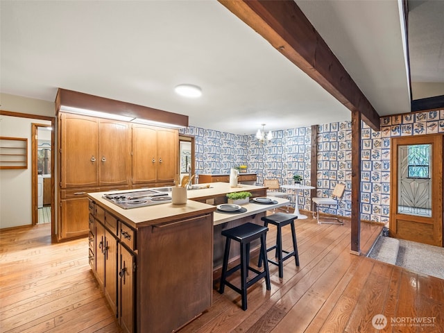 kitchen with a chandelier, light wood-style floors, beam ceiling, an island with sink, and wallpapered walls