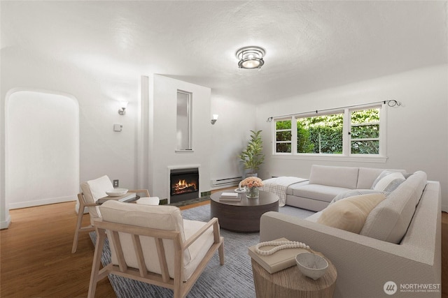 living room with a warm lit fireplace, a textured ceiling, baseboard heating, and wood finished floors