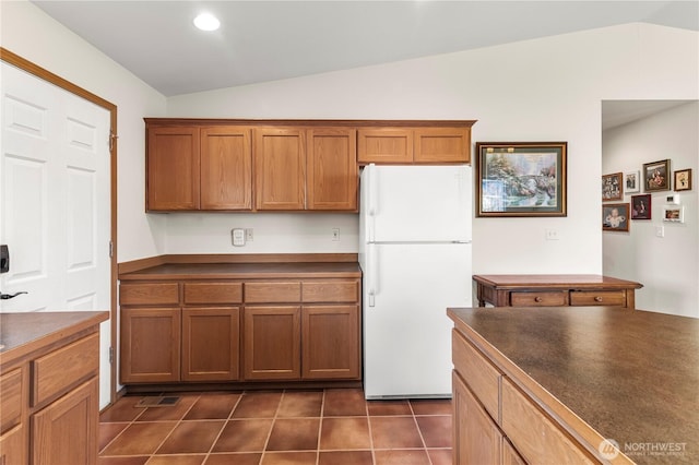 kitchen featuring brown cabinetry, dark tile patterned flooring, freestanding refrigerator, vaulted ceiling, and dark countertops