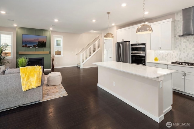 kitchen with dark wood-style floors, a glass covered fireplace, a center island, stainless steel appliances, and wall chimney range hood