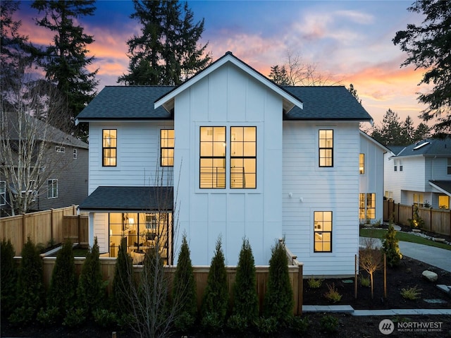 back of property at dusk featuring board and batten siding, roof with shingles, and fence