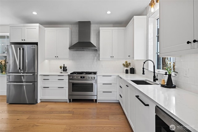 kitchen featuring light wood finished floors, wall chimney exhaust hood, appliances with stainless steel finishes, white cabinetry, and a sink