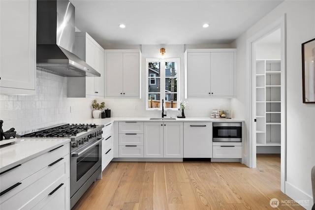 kitchen with a sink, white cabinetry, light wood-style floors, high end stainless steel range, and wall chimney exhaust hood