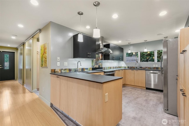 kitchen featuring a peninsula, a sink, light brown cabinetry, stainless steel appliances, and wall chimney range hood