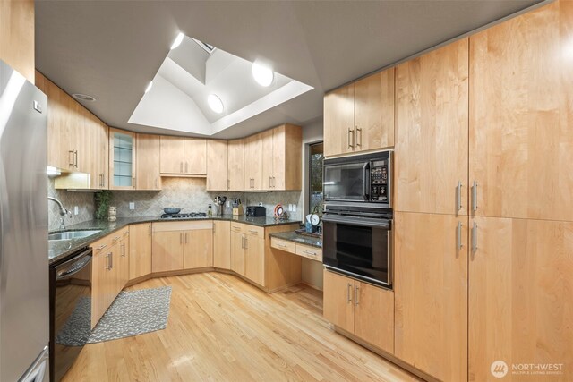 kitchen featuring a raised ceiling, light brown cabinets, a sink, dark stone countertops, and black appliances