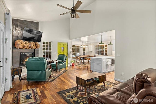 living area featuring a fireplace, ceiling fan, wood finished floors, high vaulted ceiling, and baseboards