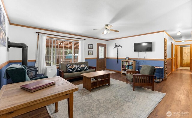 living area featuring a wainscoted wall, wood finished floors, crown molding, ceiling fan, and a wood stove