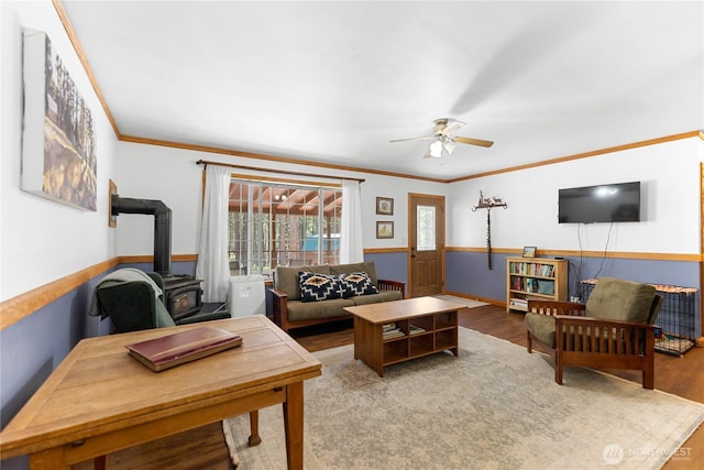 living area with a ceiling fan, wood finished floors, wainscoting, crown molding, and a wood stove