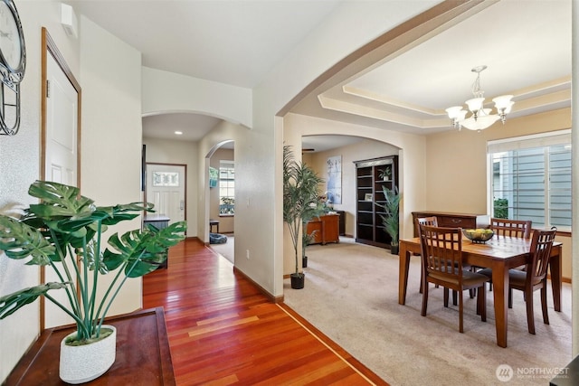 carpeted dining room with wood finished floors, baseboards, arched walkways, a raised ceiling, and a notable chandelier