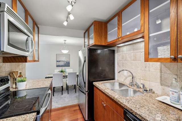 kitchen with stainless steel appliances, brown cabinetry, glass insert cabinets, a sink, and wood finished floors