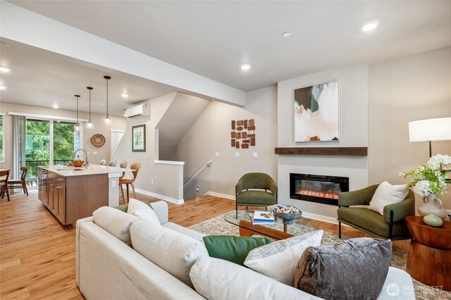 living room featuring a wall unit AC, recessed lighting, light wood-style floors, a glass covered fireplace, and baseboards