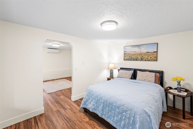 bedroom featuring arched walkways, a textured ceiling, wood finished floors, baseboards, and an AC wall unit