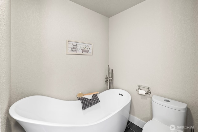 bathroom with a soaking tub, a textured wall, toilet, baseboards, and tile patterned floors