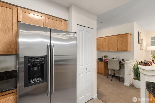 kitchen with baseboards, brown cabinetry, light colored carpet, built in study area, and stainless steel fridge with ice dispenser