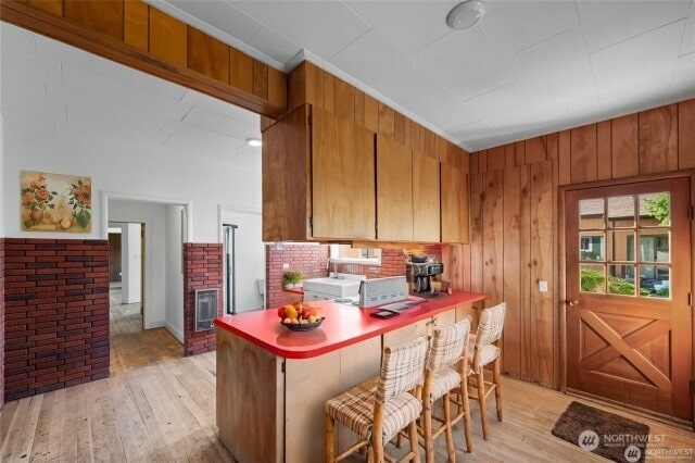 kitchen with light wood-style flooring, wooden walls, a peninsula, brown cabinets, and a kitchen bar