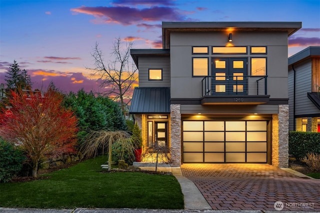 contemporary house featuring decorative driveway, an attached garage, metal roof, a balcony, and stone siding