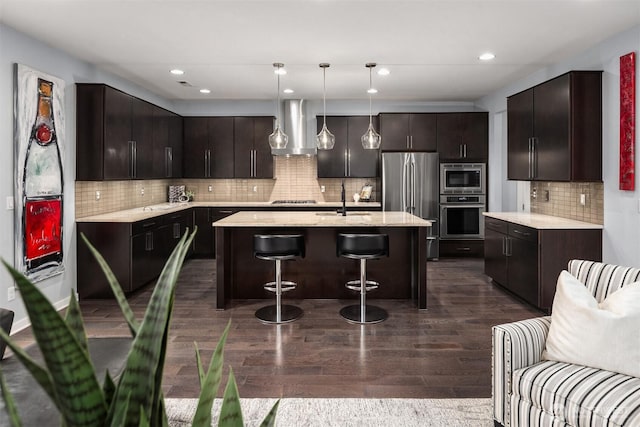 kitchen with stainless steel appliances, dark wood-type flooring, a kitchen island with sink, a sink, and wall chimney range hood