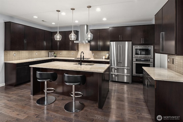 kitchen with a center island with sink, appliances with stainless steel finishes, dark wood-style flooring, light stone countertops, and exhaust hood