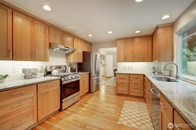 kitchen with light wood finished floors, appliances with stainless steel finishes, light stone countertops, under cabinet range hood, and a sink