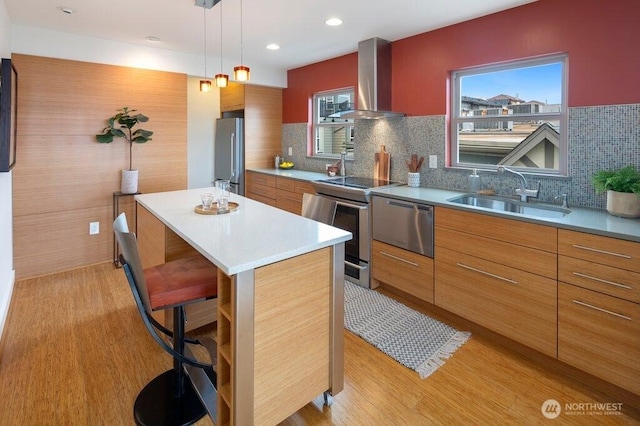 kitchen with stainless steel appliances, a kitchen island, a sink, light wood-style floors, and wall chimney range hood