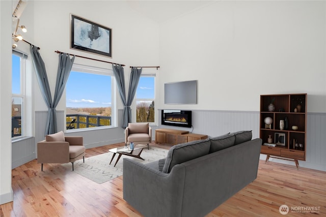 living area with light wood-style floors, a glass covered fireplace, a wainscoted wall, and a towering ceiling