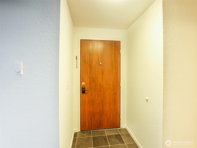 doorway with a textured wall, dark tile patterned flooring, and baseboards