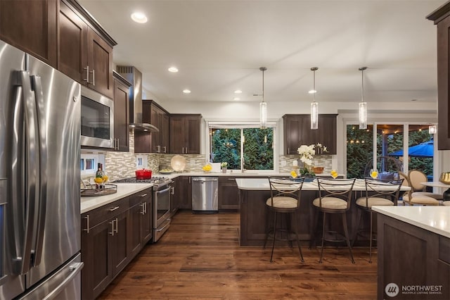 kitchen with wall chimney range hood, dark brown cabinetry, appliances with stainless steel finishes, light countertops, and dark wood-style flooring