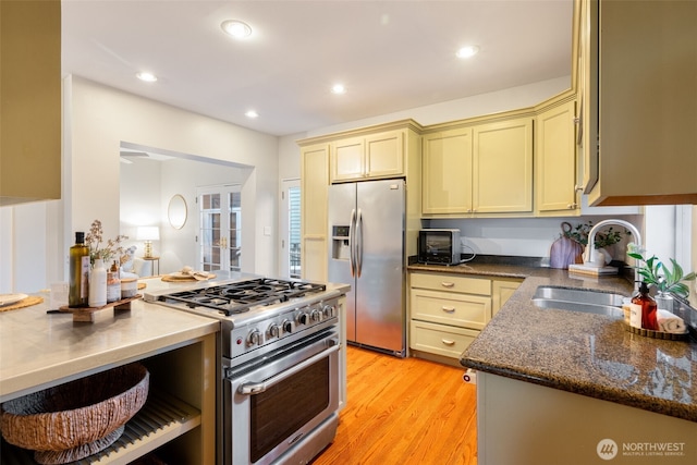 kitchen featuring light wood finished floors, cream cabinets, appliances with stainless steel finishes, a sink, and dark stone counters