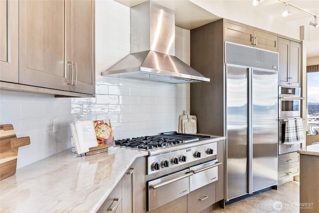 kitchen featuring wall chimney range hood, appliances with stainless steel finishes, decorative backsplash, light stone countertops, and a warming drawer