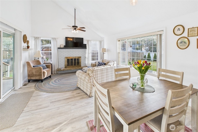 dining area featuring high vaulted ceiling, a brick fireplace, a ceiling fan, and light wood-style floors