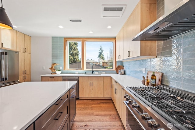 kitchen with premium appliances, a sink, light countertops, light brown cabinetry, and wall chimney exhaust hood