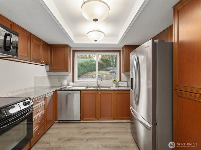 kitchen with a sink, light wood-type flooring, black appliances, a tray ceiling, and brown cabinetry