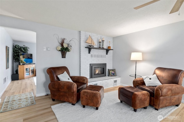 living room with baseboards, light wood-type flooring, a fireplace, a textured ceiling, and a ceiling fan