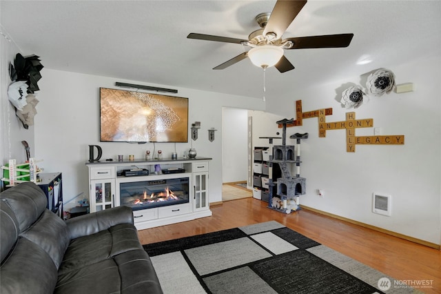 living area with baseboards, visible vents, a ceiling fan, a glass covered fireplace, and wood finished floors