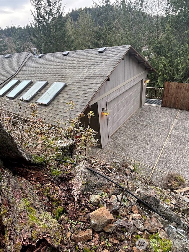 view of side of home featuring a garage, a view of trees, roof with shingles, and fence