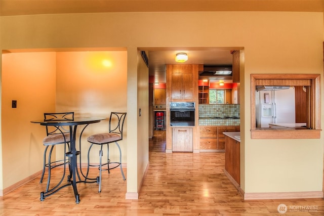 kitchen with light wood-style flooring, decorative backsplash, brown cabinetry, white fridge with ice dispenser, and black oven