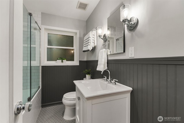 bathroom with visible vents, toilet, shower / bath combination with glass door, wainscoting, and vanity