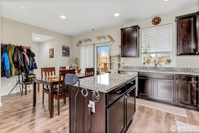 kitchen with light wood finished floors, recessed lighting, dark brown cabinetry, a kitchen island, and light stone countertops