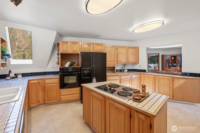 kitchen featuring tile counters, a kitchen island, black appliances, light brown cabinets, and a sink